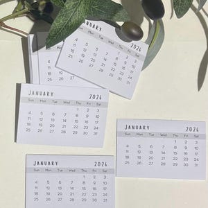 May include: Small white 2026 calendars showing January. The calendars are rectangular and display the days of the week and dates. Green leaves and olives are in the background.