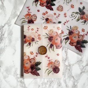 May include: A white envelope with a floral design featuring burgundy and orange flowers. The envelope has a gold wax seal with the word "Sealed" on it.
