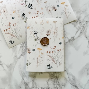 May include: A white envelope with a floral pattern and a wax seal. The envelope is folded in half and has a brown wax seal with a bee design. The envelope is on a white marble surface.