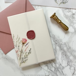 May include: A white invitation card with a pink wax seal and a floral design. The card is on top of a blush pink envelope. The card is on a white marble surface with a gold wax seal stamp.