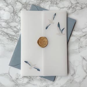 May include: A white invitation with three watercolor seagulls in flight, sealed with a gold wax stamp. The invitation is layered over a blue envelope, set against a marble background.
