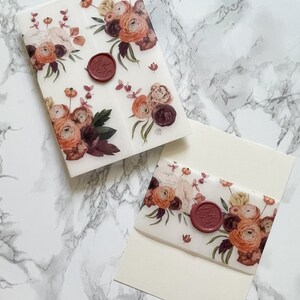 May include: Two white envelopes with a floral design and a red wax seal. The floral design features burgundy, peach, and white flowers. The envelopes are on a white marble surface.