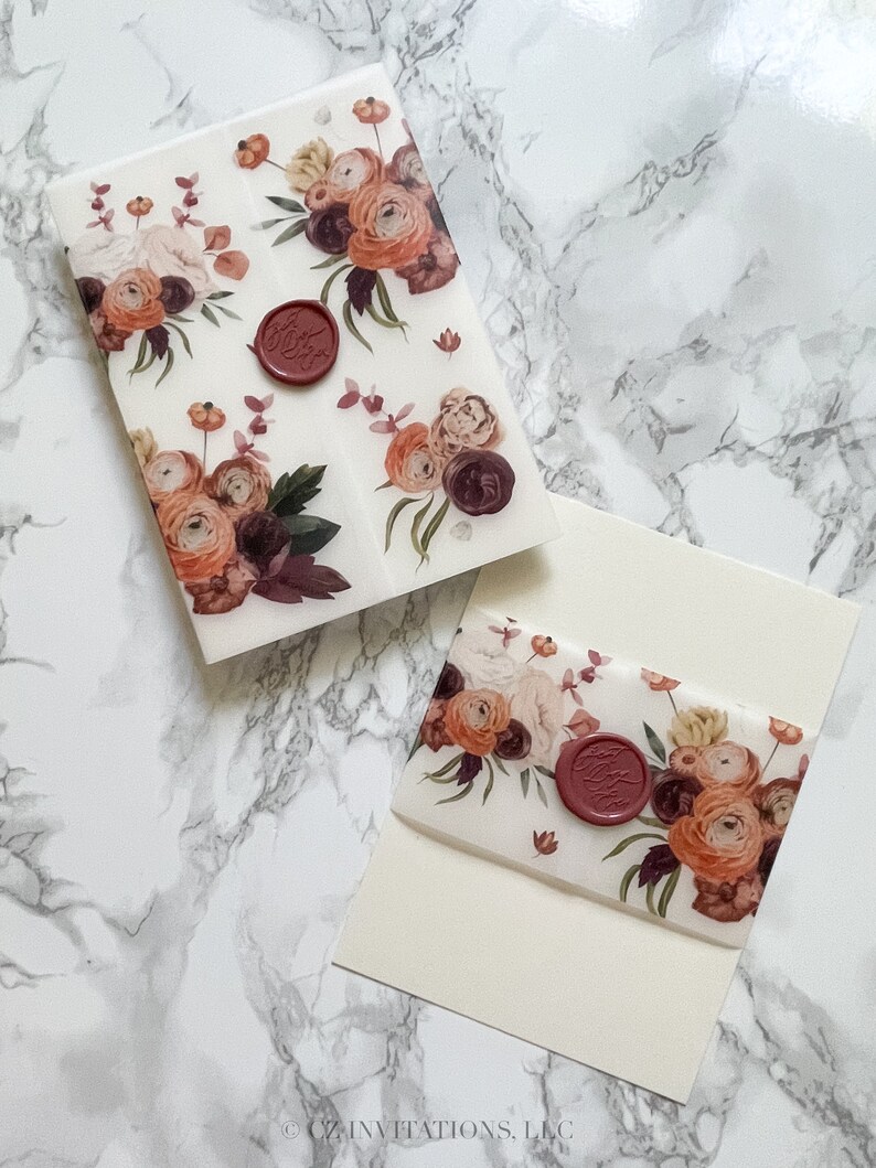 May include: Two white envelopes with a floral design and a red wax seal. The floral design features burgundy, peach, and white flowers. The envelopes are on a white marble surface.