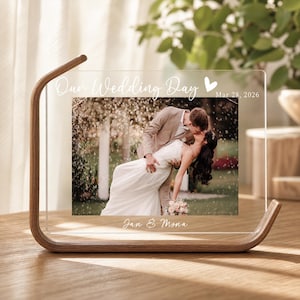 Peut inclure: Un cadre photo rectangulaire en acrylique transparent maintenu par un support en bois incurvé. Le cadre présente le texte imprimé en blanc Our Wedding Day, un cœur, la date Mar 28, 2026, et les noms Jan & Mona, entourant une photo de couple.