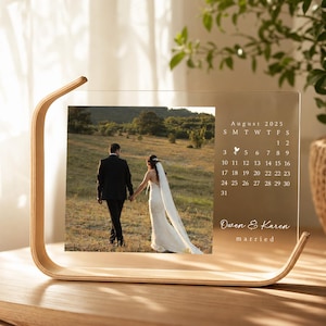 Può includere: Una cornice calendario in acrilico trasparente con base in legno chiaro che mostra la fotografia di una coppia che cammina in un campo. Il calendario mostra agosto 2025 con un'icona a forma di cuore sul 4 e il testo "Owen & Karen" seguito da "married".
