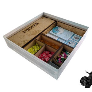 May include: A square board game box organizer with wooden dividers. The box contains a long wooden lid labeled FINSPAN, stacks of blue game cards, and four compartments filled with small yellow, orange, pink, and multi-colored game tokens.