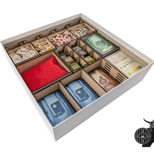 Puede incluir: Una caja de juego blanca con divisores de madera, llena de componentes del juego. La caja contiene cartas, fichas y una bolsa roja. Las piezas del juego están organizadas en compartimentos separados, con varios colores y diseños.