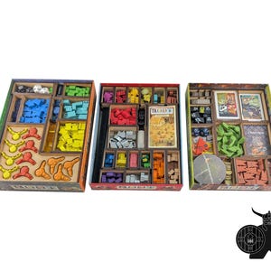 Puede incluir: Tres cajas rectangulares de juegos de mesa con insertos de madera que contienen componentes organizados. Las cajas contienen fichas de madera de varios colores, cartas, dados y marcadores. La caja central presenta una tarjeta con el texto ROOT.