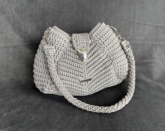 May include: A crocheted gray handbag with a silver clasp and a braided strap. The bag has a rectangular metal tag near the bottom. The bag is on a gray surface.