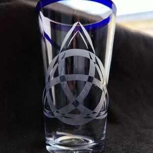 May include: A clear glass shot glass with a blue rim and an etched Celtic knot design.