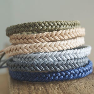 May include: Five braided bracelets in different colors, including green, beige, light blue, and dark blue. The bracelets are stacked on top of each other.