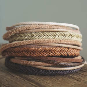 Könnte beinhalten: Ein Stapel geflochtener Lederarmbänder in verschiedenen Brauntönen, Beige und Schwarz. Die Armbänder sind auf einer Holzoberfläche gestapelt.