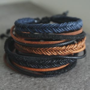 May include: A collection of braided and leather bracelets in shades of navy, black, and brown. Some bracelets feature a woven design, while others are made of smooth leather. The bracelets are stacked together.