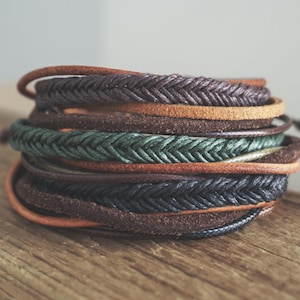 May include: A collection of leather bracelets, some braided, in shades of brown, green, and black. The bracelets are displayed on a wooden surface, highlighting the different textures and colours of the leather.