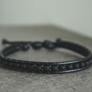 Könnte beinhalten: Ein schwarzes Armband mit Perlen und einem geflochtenen Lederband. Das Armband hat ein doppelt gewickeltes Design mit einer Reihe von mattschwarzen Perlen. Das Armband ist verstellbar und hat einen Knotenverschluss.