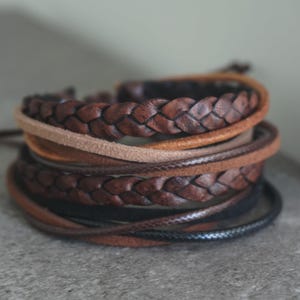 May include: A collection of leather bracelets in shades of brown and black. The bracelets feature braided and smooth designs, creating a layered effect. The textures and colours suggest a rustic, handcrafted aesthetic.