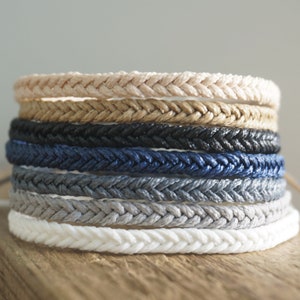 May include: A stack of eight braided rope bracelets in various colours, including beige, brown, black, navy blue, grey, and white. The bracelets are stacked on a wooden surface.
