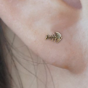 May include: A gold fish bone shaped earring in a person's earlobe.