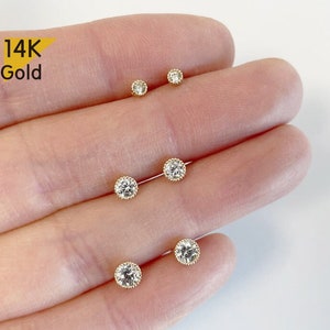 May include: Five 14K gold earrings with small round clear stones. The earrings are arranged in a line on a hand.