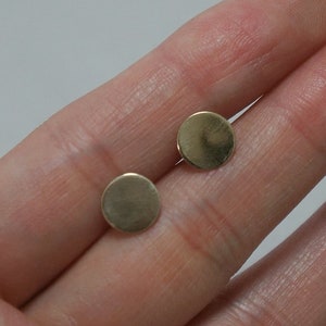May include: Two small, round, gold-colored stud earrings. The earrings are simple and minimalist in design, with a flat, disc-like shape. The earrings are held in a person's hand.