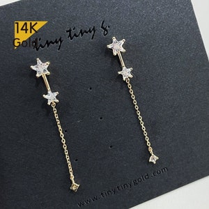 Puede incluir: Un par de pendientes de oro de 14 quilates con un colgante de estrella. Los pendientes tienen dos estrellas pequeñas y una cadena más larga con una sola estrella al final. Los pendientes se muestran en una tarjeta negra con el texto "14K Gold Tiny & www.tinytinygold.com".