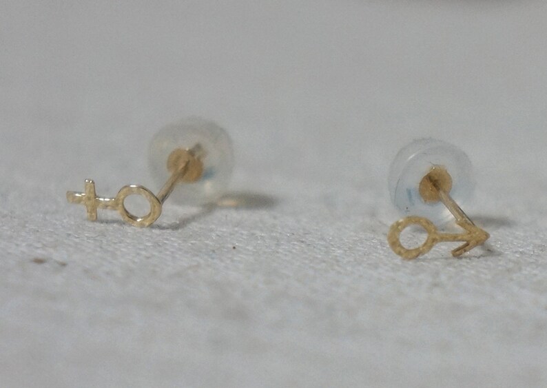 Puede incluir: Un par de pendientes de oro con un dise&ntilde;o sencillo. Un pendiente presenta un c&iacute;rculo con una cruz encima, mientras que el otro pendiente presenta un c&iacute;rculo con una peque&ntilde;a flecha apuntando hacia la derecha.