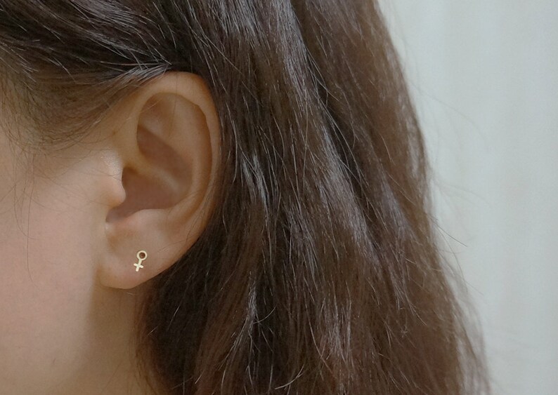 Puede incluir: Primer plano de la oreja de una persona con un peque&ntilde;o pendiente de oro en forma de s&iacute;mbolo femenino.