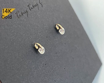 May include: A pair of small, gold earrings with a single clear stone on each earring. The earrings are on a black background with the text "14K Gold" in gold lettering.