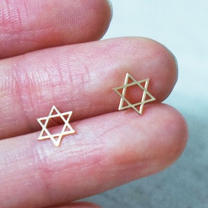 Pode incluir: Dois pequenos brincos dourados em forma de Estrela de David. Os brincos têm a forma de uma estrela de seis pontas, com um design simples e minimalista. São mostrados num fundo claro.