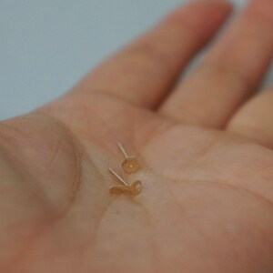 May include: Two gold-toned earrings, one shaped like a hexagon and the other shaped like a wrench, are displayed on a person's hand.