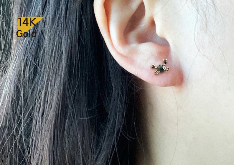 May include: 14K Gold crab earrings with a black gemstone. The earrings are shown in a person's ear.