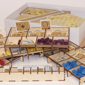 May include: A wooden organiser for a board game with multiple compartments for storing game pieces, cards, and tiles. The organiser is made of light brown wood and has a white label with the text "Hospital" on it.