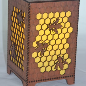 May include: A wooden lamp shaped like a honeycomb with a yellow light shining through. The lamp has four bees cut out of the wood.