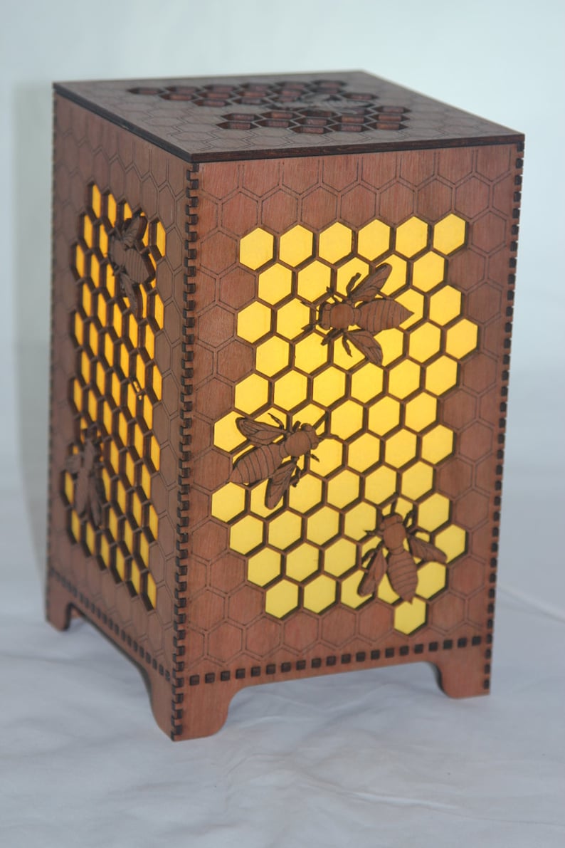 May include: A wooden lamp shaped like a honeycomb with a yellow light shining through. The lamp has four bees cut out of the wood.