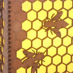 May include: A wooden honeycomb lampshade with two bees cut out of the wood. The honeycomb is illuminated from behind, creating a warm glow.