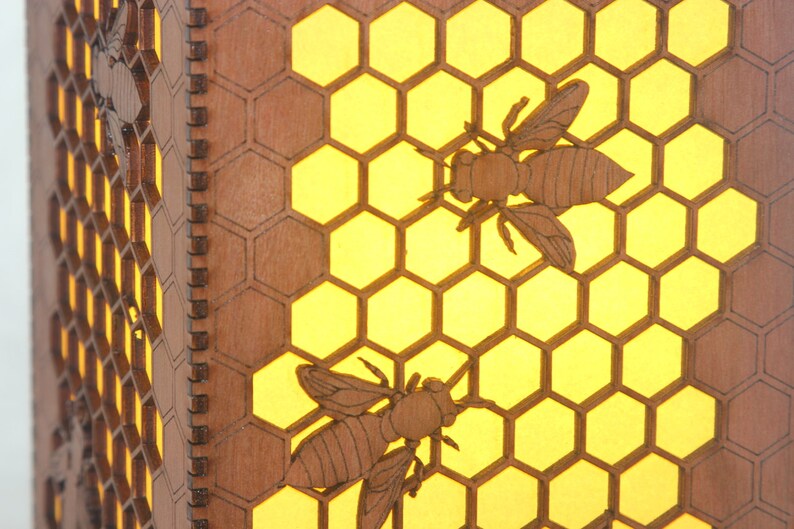 May include: A wooden honeycomb lampshade with two bees cut out of the wood. The honeycomb is illuminated from behind, creating a warm glow.