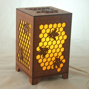 May include: A wooden lamp with a honeycomb design and bee silhouettes. The lamp is lit and casts a warm glow.