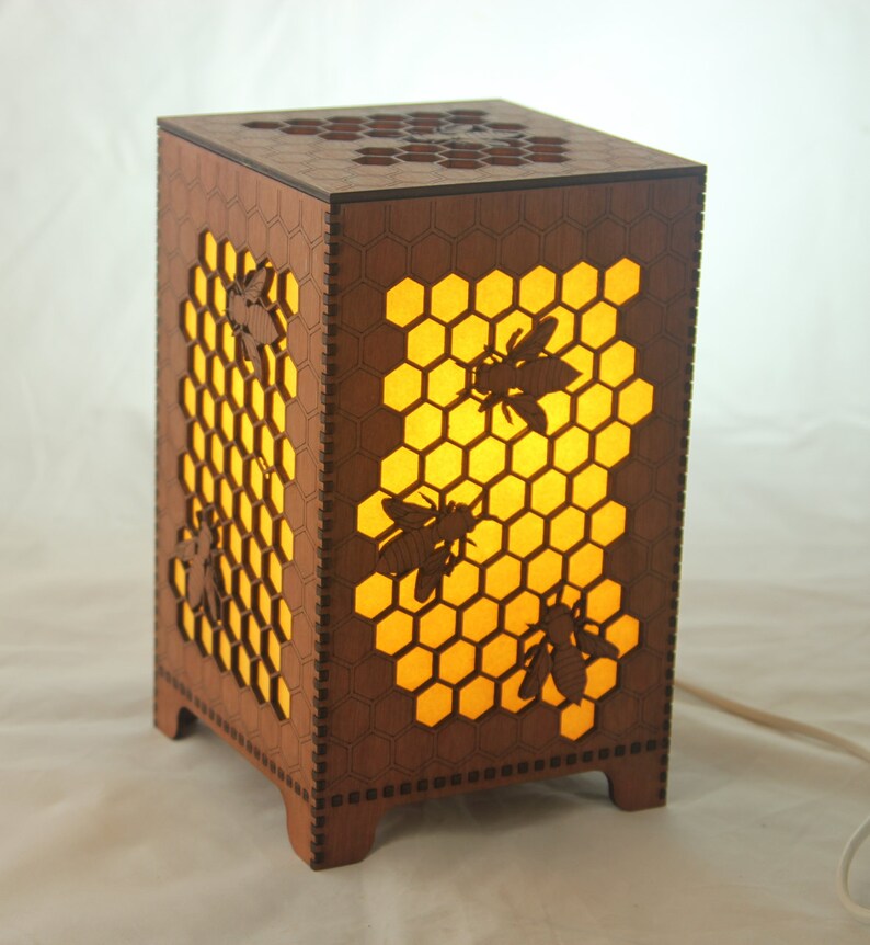 May include: A wooden lamp with a honeycomb design and bee silhouettes. The lamp is lit and casts a warm glow.