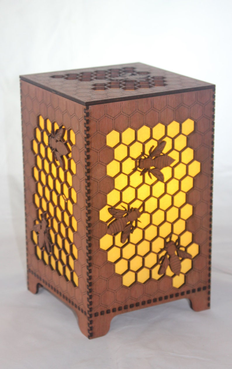 May include: A wooden lamp with a honeycomb design and cut-out bees. The lamp is lit and the honeycomb glows yellow.