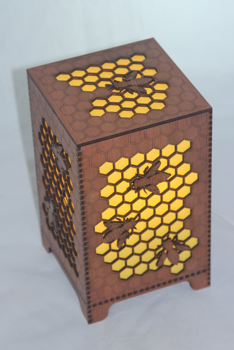 May include: A wooden lamp with a honeycomb design and bees cut out. The lamp is lit from the inside, casting a warm glow.