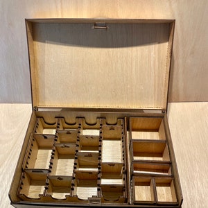 Puede incluir: Una caja de madera con tapa abatible y varios compartimentos para organizar piezas de juego. La caja est&aacute; hecha de madera marr&oacute;n claro y tiene un acabado natural.