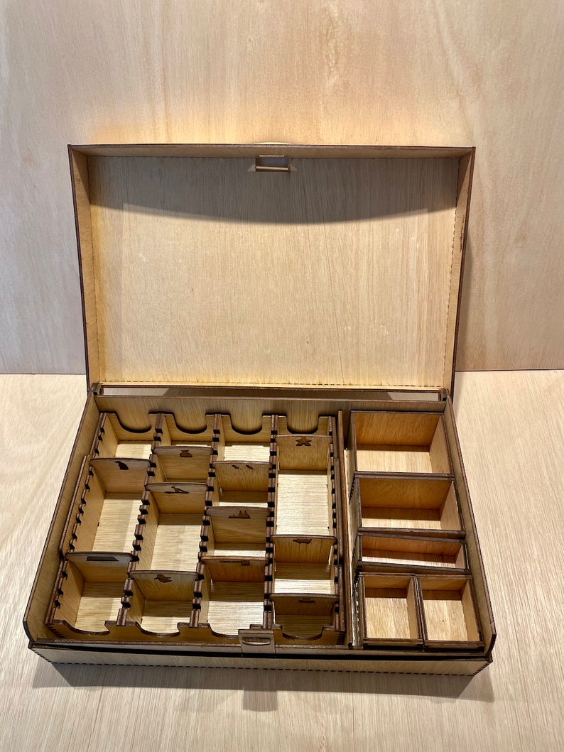 Puede incluir: Una caja de madera con tapa abatible y varios compartimentos para organizar piezas de juego. La caja est&aacute; hecha de madera marr&oacute;n claro y tiene un acabado natural.