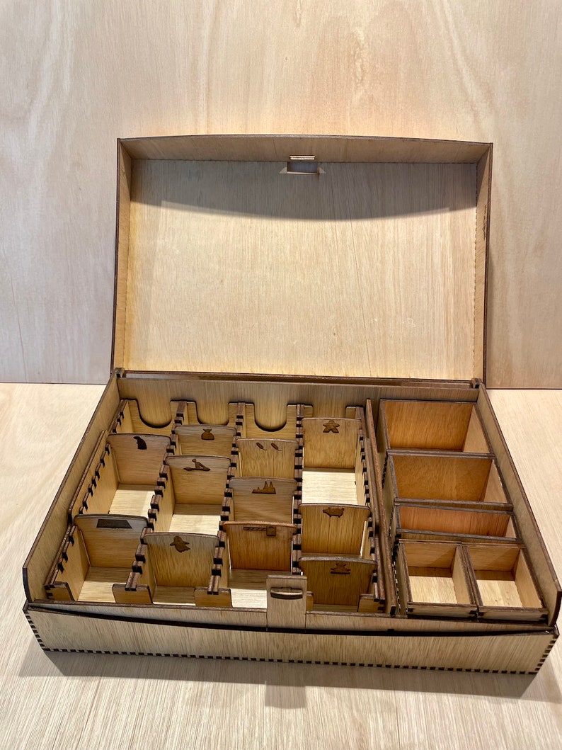 Puede incluir: Una caja de madera con tapa abatible y m&uacute;ltiples compartimentos para organizar piezas de juego. La caja est&aacute; hecha de madera marr&oacute;n claro y tiene un acabado natural.