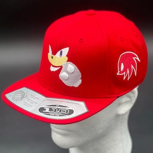 May include: A red baseball cap featuring an embroidered image of a cartoon character on the front and side. The cap has a flat brim and the text "110 ONE TEN" on the brim.
