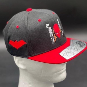 May include: Black and red baseball cap with a flat brim. The cap features a red bat symbol on the side and a graphic of a red helmet and guns. The brim is red, and the top of the cap has red accents.