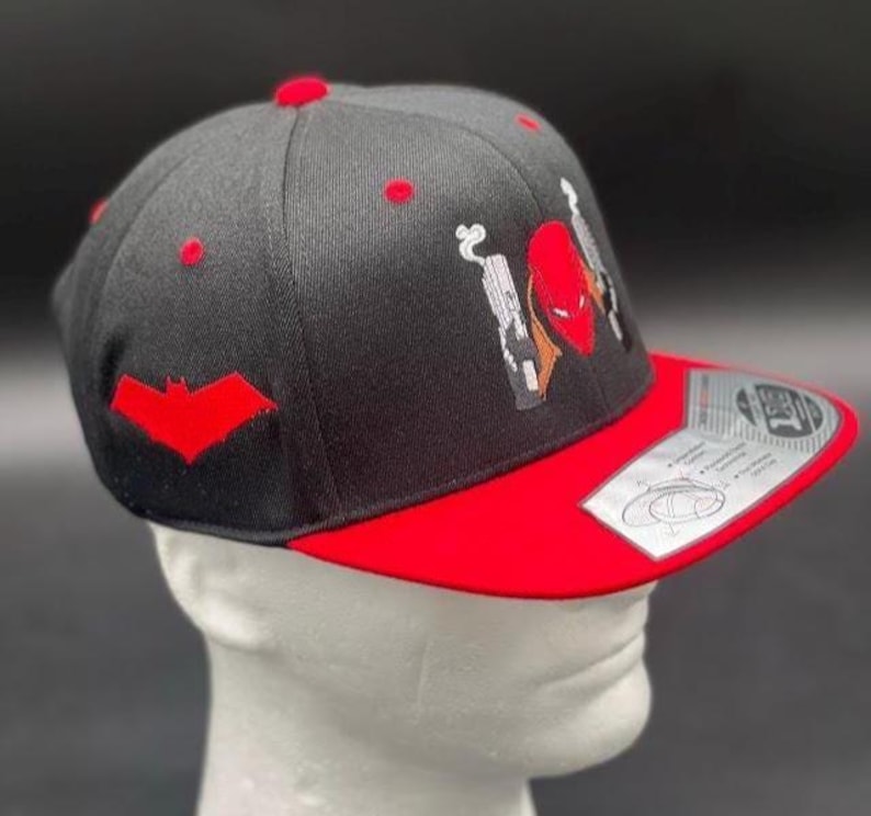 May include: Black and red baseball cap with a flat brim. The cap features a red bat symbol on the side and a graphic of a red helmet and guns. The brim is red, and the top of the cap has red accents.