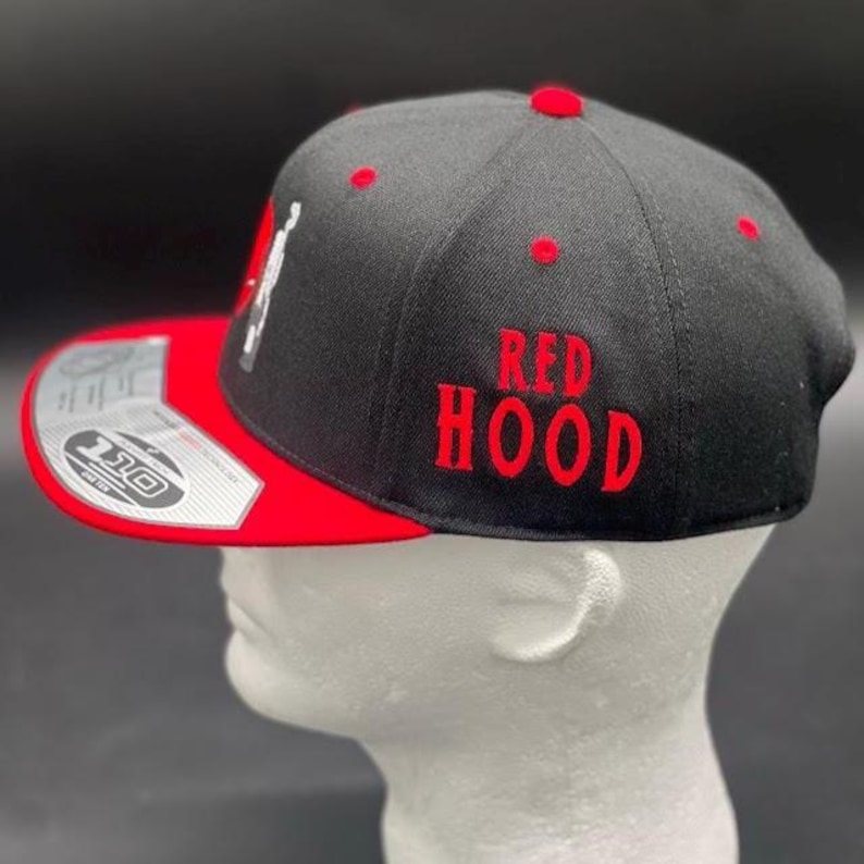 May include: Black baseball cap with a red brim and red embroidered text that reads "RED HOOD". The cap has red accents and a white graphic on the side. The cap is displayed on a white mannequin head.