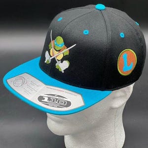 May include: Black and blue baseball cap featuring an embroidered Teenage Mutant Ninja Turtle character with crossed swords. The cap has a blue brim and a blue button on top. The side of the cap has a patch with the letter "L".