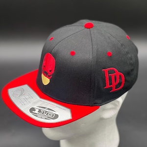 May include: Black and red baseball cap with a red embroidered skull and the letter "D" on the front. The cap has a red underside and a "Flexfit 110" logo.