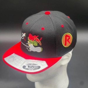 May include: Black and red baseball cap featuring an embroidered Teenage Mutant Ninja Turtle design. The cap has a red bill and a red button on top. The cap has the number "110" on the bill.